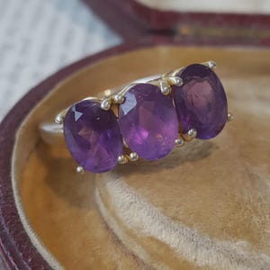 May include: A three-stone ring featuring oval-cut purple amethyst gemstones set in a polished silver-toned metal band. The stones are arranged horizontally, each secured by four small metal prongs, creating a classic and symmetrical jewellery design.