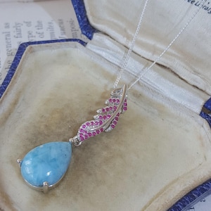 Peut inclure: Un collier en argent avec un pendentif en pierre bleu clair en forme de goutte attaché à une breloque en forme de plume incurvée. La plume est incrustée de petites pierres roses vives et d'accents clairs, suspendue à une fine chaîne en argent.