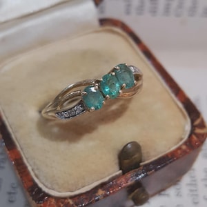 May include: A gold ring featuring three oval-cut green gemstones set in a row, flanked by small clear stones on each side of the band. The ring has a split shank design and is presented in a beige velvet-lined box with a brown patterned exterior.