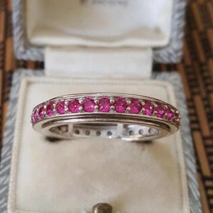 May include: A silver-toned band ring featuring a continuous row of small, round, vibrant pink stones set along the centre. The metal band has a polished finish with a smooth, raised border framing the stones, creating a sleek and uniform appearance.