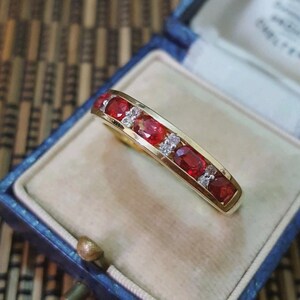 May include: A gold band ring featuring a row of alternating oval-cut red rubies and small clear round-cut diamonds set in a channel setting. The ring is displayed inside a blue velvet-lined jewelry box with a cream-colored interior cushion.