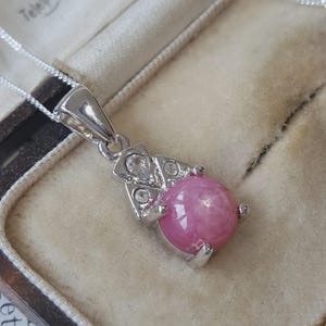 Könnte beinhalten: Eine silberfarbene Anhängerkette mit einem runden, im Cabochon-Schliff gearbeiteten rosa Stein, der eine sternförmige Lichtreflexion aufweist. Der Anhänger ist mit drei kleinen, klaren, rund geschliffenen Steinen in einer silberfarbenen Fassung verziert.