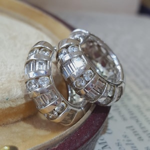 Op de afbeelding: Een paar zilverkleurige oorringen met een kanaalzetting. Elke ring is versierd met afwisselende rijen van heldere, rond geslepen stenen en rechthoekige baguette-geslepen stenen, wat zorgt voor een reflecterend metalen oppervlak met heldere accenten.