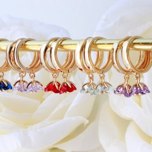 May include: A set of eight gold hoop earrings with dangling charms. Each charm features a cluster of three sparkling crystals in different colors, including clear, blue, pink, red, green, purple, and champagne.