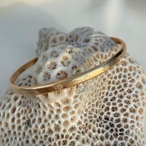 May include: A gold-colored bangle bracelet with star-shaped designs. The bracelet is resting on a textured, off-white surface. The stars are evenly spaced around the bracelet. The bangle is a simple, elegant piece of jewelry.