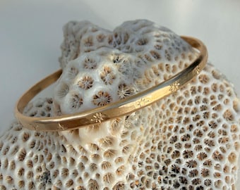 May include: A gold-colored bangle bracelet with star-shaped engravings. The bracelet is resting on a textured, off-white surface. The bangle is a simple, elegant piece of jewelry.