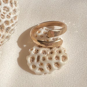 May include: A gold-toned hammered metal ring with a unique twisted design. The ring is shown on a white coral-like surface.