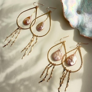 May include: A pair of gold hoop earrings with a teardrop-shaped pearl pendant and dangling chains. The pearls are a soft pink and white color.