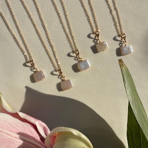 May include: Several gold-toned necklaces with rectangular, iridescent pendants are displayed. The necklaces are arranged on a light-colored surface, with a pink and white flower and green leaf in the foreground. The pendants appear to be made of pearl.