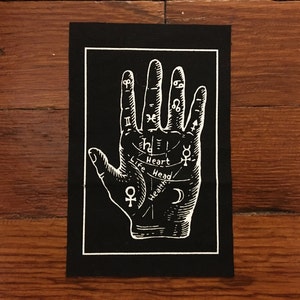 Pode incluir: Um remendo de tecido preto com um contorno branco que representa uma mão com símbolos astrológicos e linhas. A mão está etiquetada com "Life", "Head", "Heart" e "Health".