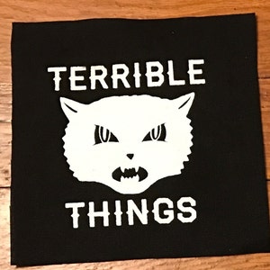 Pode incluir: Remendo preto com texto branco que diz "TERRIBLE THINGS" e um gato de desenho animado branco com olhos zangados e uma boca grande.
