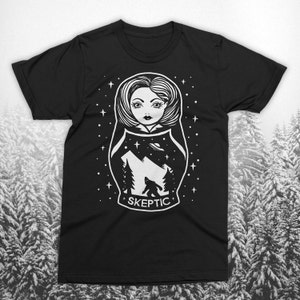 May include: Black t-shirt featuring a white graphic of a Matryoshka doll with a mountain scene, Bigfoot, and a UFO. The word "SKEPTIC" is below the graphic. The design includes stars and trees.