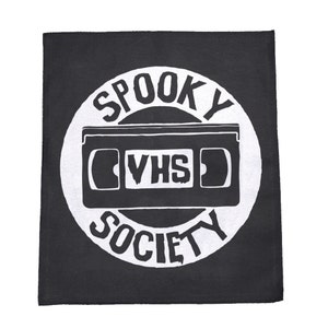 May include: Black and white patch with a circle design. The circle has the text "Spooky VHS Society" around the edge. Inside the circle is a white silhouette of a VHS tape.