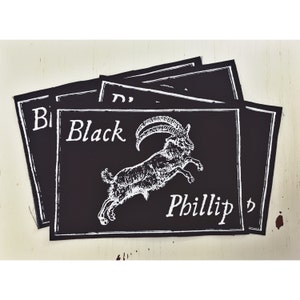 Pode incluir: Patch de ferro de passar a ferro preto e branco com uma cabra saltando e o texto "Black Phillip" em uma fonte cursiva.