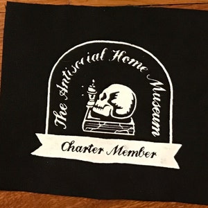 Pode incluir: Patch preto e branco com uma caveira segurando uma vela e um livro. O texto diz "The Antisocial Home Museum" e "Charter Member".
