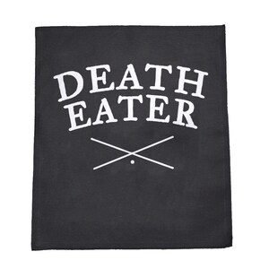 May include: Black square patch with the words "DEATH EATER" in white, arched text. Below the text are two crossed lines with a small dot at the intersection. The patch is likely made of fabric.