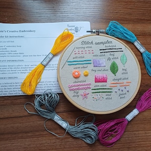 May include: Embroidery kit featuring a wooden hoop, instructions, and colourful threads. The sampler hoop showcases different stitch examples, including running and leaf stitches. Includes yellow, blue, grey, and pink thread.