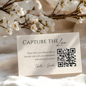 May include: A rectangular cream-colored card featuring the black text CAPTURE THE Love, instructions to share photos via a QR code, and the names Sophia + Jordan. The card has a textured surface and is positioned against white flowers.