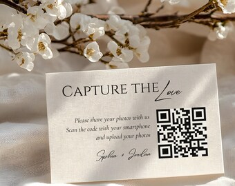 Può includere: Un biglietto color crema con la scritta "CAPTURE THE Love" e un codice QR. Il biglietto include istruzioni per condividere foto tramite smartphone. Delicati fiori e rami bianchi sullo sfondo. I nomi "Sophia + Jordan" sono in basso.