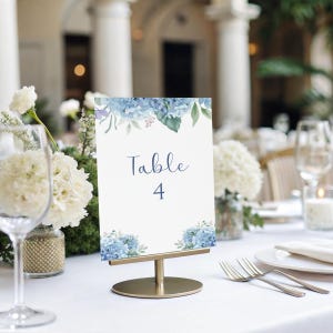May include: A white table number card with blue floral accents and the text "Table 4." The card is held in a gold metal stand. The table is set with white linens, flowers, and glassware, suggesting a wedding or event.