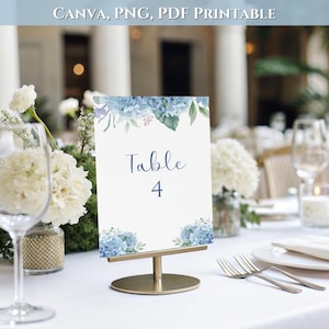 May include: A vertical rectangular table sign featuring blue hydrangea floral illustrations at the top and bottom corners. The center displays the dark blue text "Table 4". The sign is held upright by a gold-toned metal stand on a white surface.