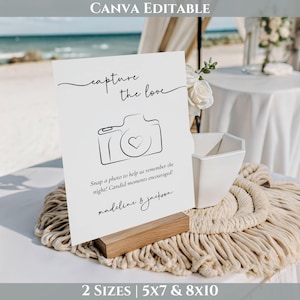 May include: A white wedding sign with the text "capture the love" and a camera illustration. The sign includes the names "madeline & jackson" and the text "Snap a photo to help us remember the night! Candid moments encouraged!" The sign is on a wooden base.