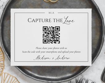 Può includere: Un biglietto d'invito a nozze con la scritta "CAPTURE THE Love" e un codice QR. Il biglietto è su un piatto grigio scuro, con due fedi d'oro sopra. I nomi "Madison + Andrew" sono scritti in basso.