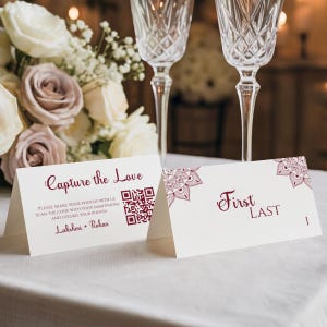 以下が含まれることがあります： バーガンディ色のテキストとフローラルアクセントが付いた、2つの白い結婚式のテーブルカード。1枚のカードにはQRコードとともに「Capture the Love」と書かれています。もう1枚のカードには「First Last」と数字の1が書かれています。カードは白いテーブルクロスの上にあります。