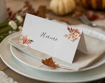 May include: A white place card with the word "Name" in elegant script. Floral arrangements in orange, brown, and cream decorate the corners. The card sits on a folded beige napkin, on a white plate, with pumpkins in the background.