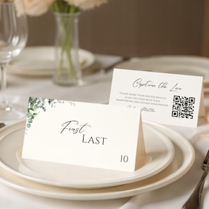 May include: Two ivory place cards with black text and a sprig of green leaves. One card reads "First Last" and "10". The other card says "Capture the Love" and includes a QR code. Wedding table setting.