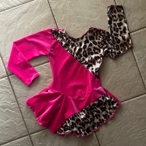 May include: A pink velvet skating dress with a leopard print design on the sleeves and skirt. The dress has a ruffled skirt and long sleeves.