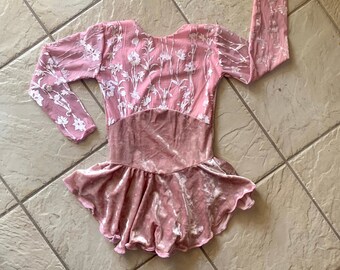 May include: A pink velvet figure skating dress with long sleeves and a white floral pattern. The dress has a fitted bodice and a flared skirt.