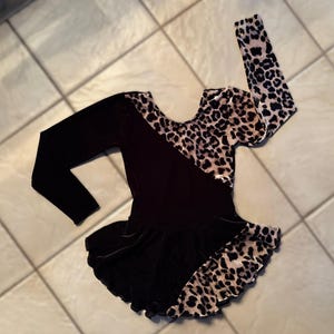 May include: A black and leopard print leotard with long sleeves. The leotard has a scoop neck and a flared skirt. The leopard print is on the sleeves and the upper bodice. The leotard is likely for dance or gymnastics.