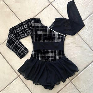 May include: Black and silver plaid long-sleeved leotard with a black skirt. The bodice features a V-neckline with pearl accents. The leotard has a black mesh panel and a black skirt.