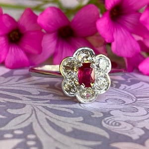 Puede incluir: Un anillo de plata con un diseño floral. El anillo presenta un rubí rectangular en el centro, rodeado por un halo de piedras preciosas redondas y claras. El anillo se encuentra sobre un fondo de flores rosas.