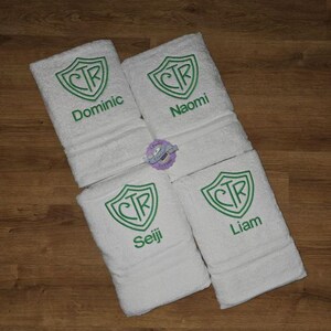 May include: Four white personalized towels with green embroidered crests and names: Dominic, Naomi, Seiji, and Liam. The towels are folded and arranged on a wooden surface.