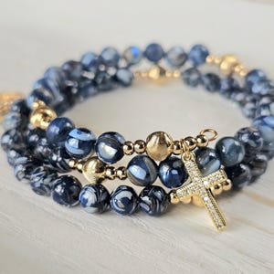 May include: Three stacked bracelets with blue and white marbled beads and gold accents. One bracelet features a gold cross pendant with clear stones. A gold charm is visible on the side.