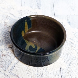 May include: A small, round ceramic bowl with a dark brown glaze and blue and yellow accents. The bowl is glazed on the inside and outside.