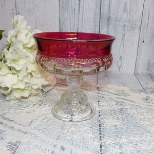 Peut inclure: Un bol en verre transparent sur un piédestal avec un bord rouge rubis et une base en verre transparent. Le bol est posé sur une nappe en dentelle blanche.