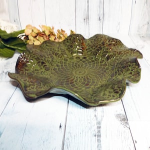 May include: A large, green ceramic serving dish with a wavy edge and a decorative, embossed pattern. The dish has a brown glaze that gives it a rustic look.