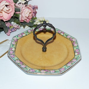 May include: An octagonal serving tray with a floral border and a central handle. The tray has a light yellow centre and a decorative edge with pink and white roses on a green background. The handle is dark metal.