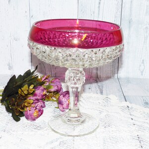 Peut inclure: Un bol en verre transparent sur un piédestal avec un motif en diamant et un bord rose foncé. Le bol est posé sur une dentelle blanche.