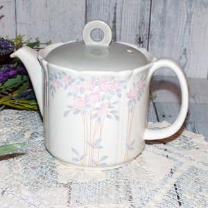 Peut inclure: Une théière en céramique blanche avec un couvercle gris et un motif floral composé de roses roses et grises. La théière a un bord festonné et une anse.