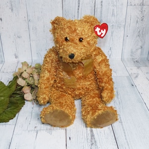May include: A golden brown teddy bear with a gold ribbon around its neck. The bear has a Ty tag attached to its ear.