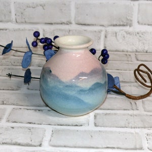 May include: A small, round ceramic vase with a light pink and blue gradient design. The vase has a white rim and a rounded body. Decorative blue berries and leaves are arranged around the vase.