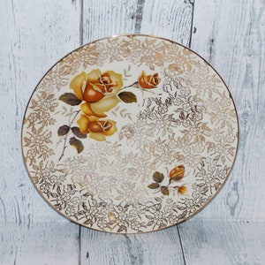 May include: A white ceramic plate with a gold and brown floral design. The plate features a large rose with a yellow centre and brown edges. The plate is decorated with a delicate floral pattern in gold.