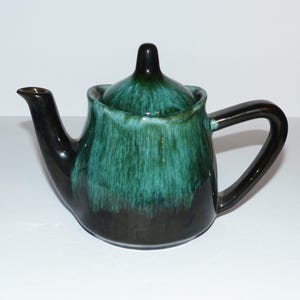 May include: A small, green and black ceramic teapot with a lid. The teapot has a rounded body and a spout on the side. The handle is on the opposite side of the spout.