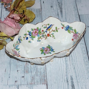 May include: A white porcelain bowl with a scalloped edge and gold trim. The bowl features a floral design with a blue bird and pink, red, and blue flowers. The bowl is on three small feet.