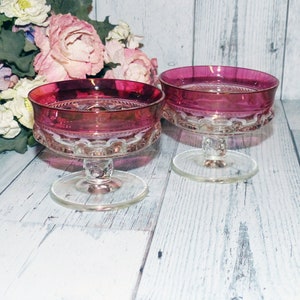 May include: Two clear glass dessert dishes with a pink rim and a scalloped edge. The dishes are on a white wooden surface.