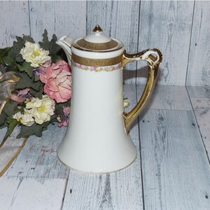 May include: A white porcelain cream jug with a gold handle and floral design. The jug has a bell-shaped body and a lid with a gold knob.